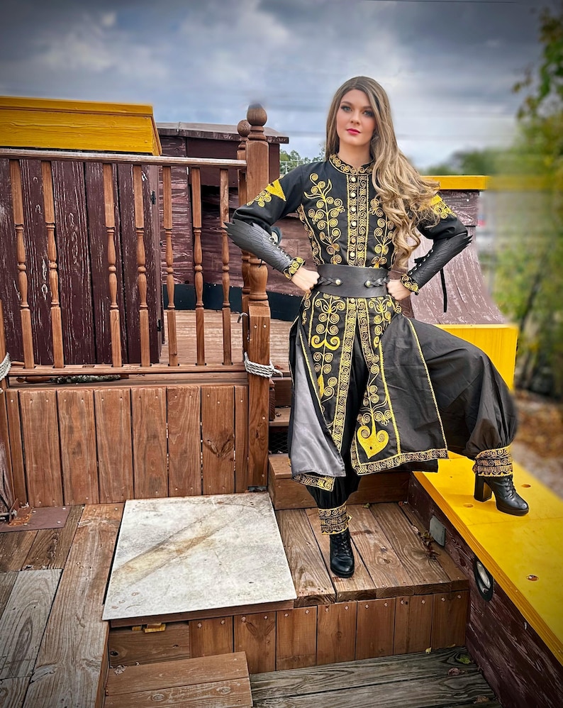 A woman wearing a black and gold embroidered costume with a long skirt and pants. She is standing on a wooden platform with a yellow railing. Elizabeth Swann from POTC.