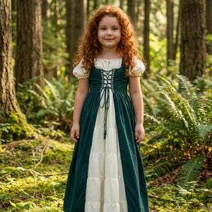 May include: A young person with long, curly red hair wearing a green and cream-colored dress with a laced bodice and short, puffed sleeves. The dress has a tiered skirt and is set against a backdrop of a lush forest with tall trees and ferns.
