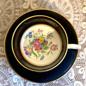 May include: A vintage teacup and saucer set. The cup and saucer are navy blue with gold trim. The inside of the cup features a floral design with pink, blue, yellow, and purple flowers. The set is placed on a white lace tablecloth.