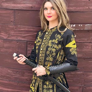 A woman wearing a black and gold embroidered jacket with a black sword. She is standing in front of a wooden wall. Elizabeth Swann costume.
