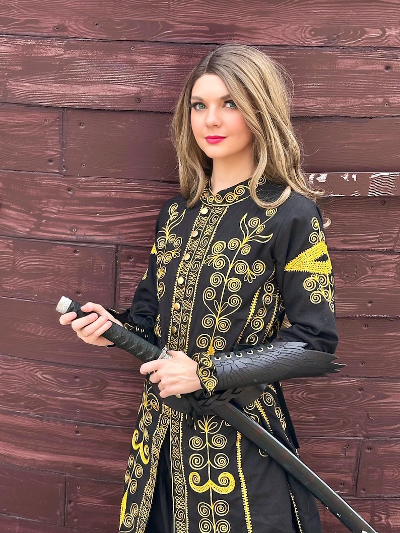 A woman wearing a black and gold embroidered jacket with a black sword. She is standing in front of a wooden wall. Elizabeth Swann costume.