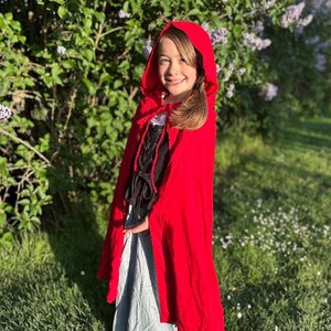 May include: A young person wearing a bright red hooded cape, black bodice with lacing, and a gray skirt. The outfit is reminiscent of a fairytale character. The background features green foliage and sunlight.