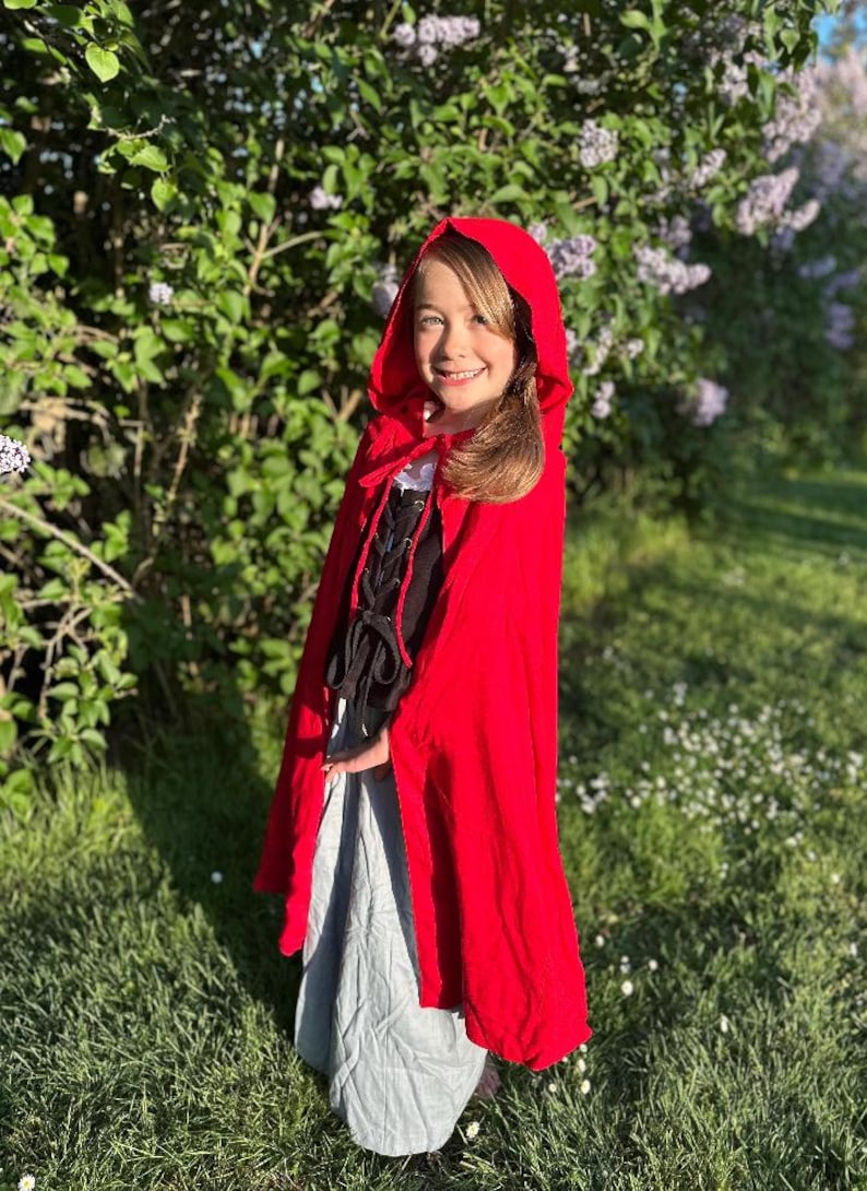 May include: A young person wearing a bright red hooded cape, black bodice with lacing, and a gray skirt. The outfit is reminiscent of a fairytale character. The background features green foliage and sunlight.