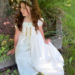 May include: A young person wearing a white dress with a floral crown. The dress features a bodice with colorful accents and a flowing skirt. The person is seated outdoors near a tree and greenery, creating a whimsical, fairytale aesthetic.