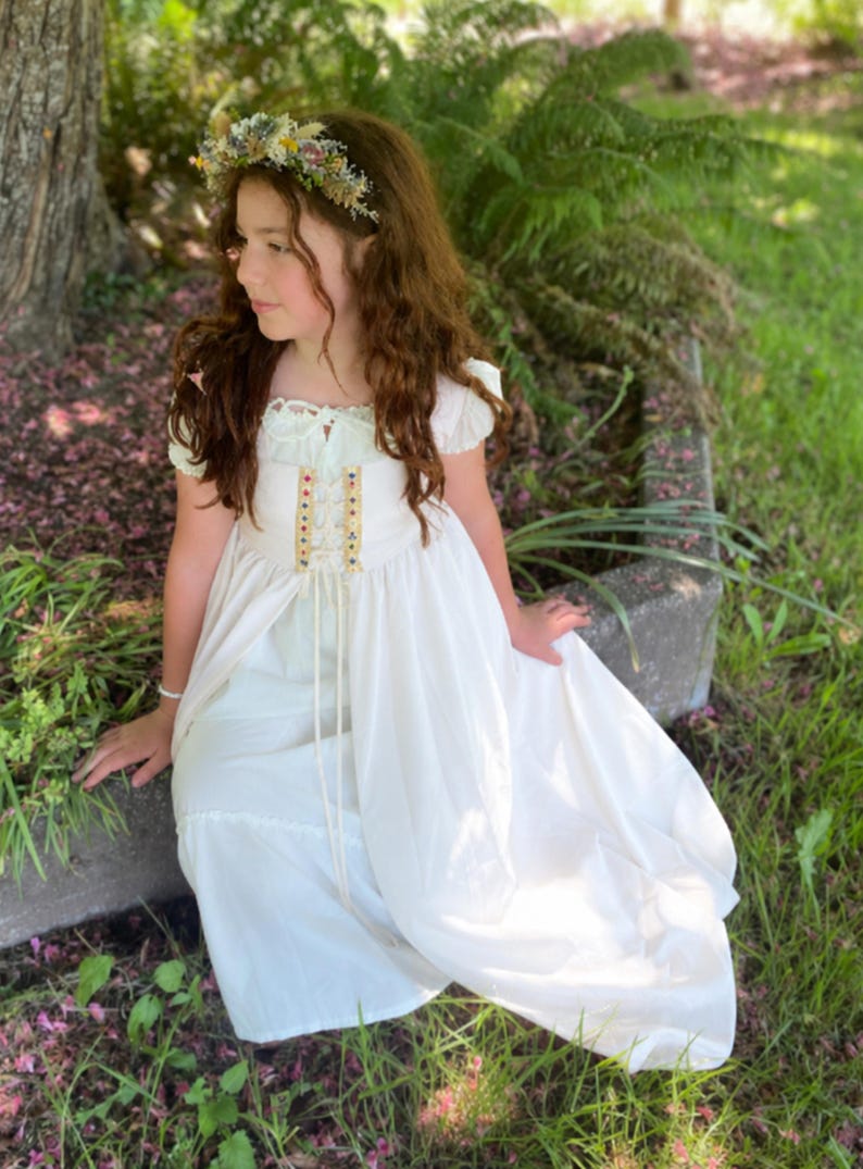 May include: A young person wearing a white dress with a floral crown. The dress features a bodice with colorful accents and a flowing skirt. The person is seated outdoors near a tree and greenery, creating a whimsical, fairytale aesthetic.