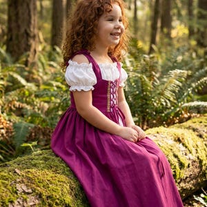 May include: A young girl wearing a magenta dress with a white off-the-shoulder blouse, sitting on a moss-covered log in a forest. The dress features a laced bodice with gold trim. The girl has curly red hair and is smiling.