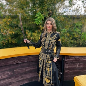 A woman in a black and gold embroidered costume holds a sword. She is standing on a wooden platform with a yellow railing. Elizabeth Swann costume