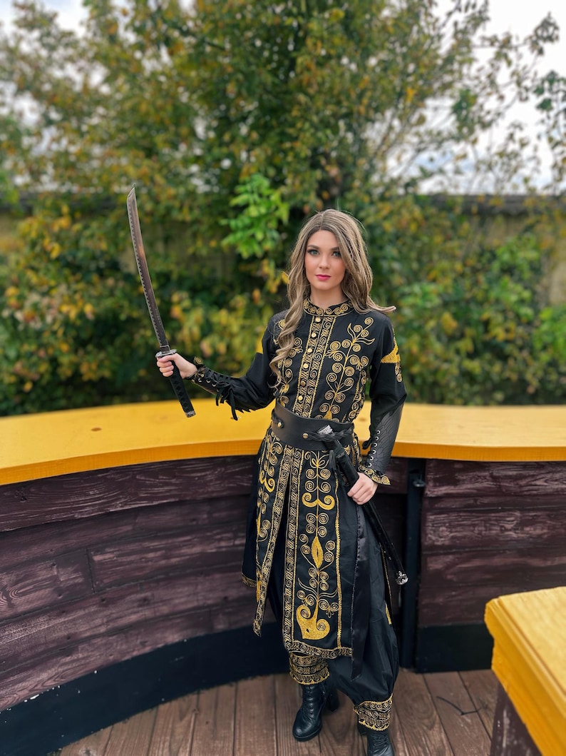 A woman in a black and gold embroidered costume holds a sword. She is standing on a wooden platform with a yellow railing. Elizabeth Swann costume