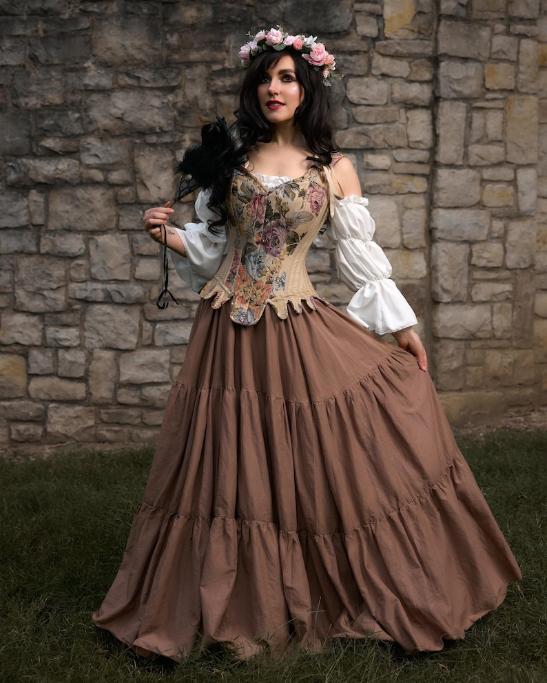 A Renaissance-style ensemble featuring a floral tapestry corset, a tiered brown skirt, and white off-the-shoulder sleeves. The outfit is complemented by a floral crown and a black feather accessory, creating a romantic, historical aesthetic.