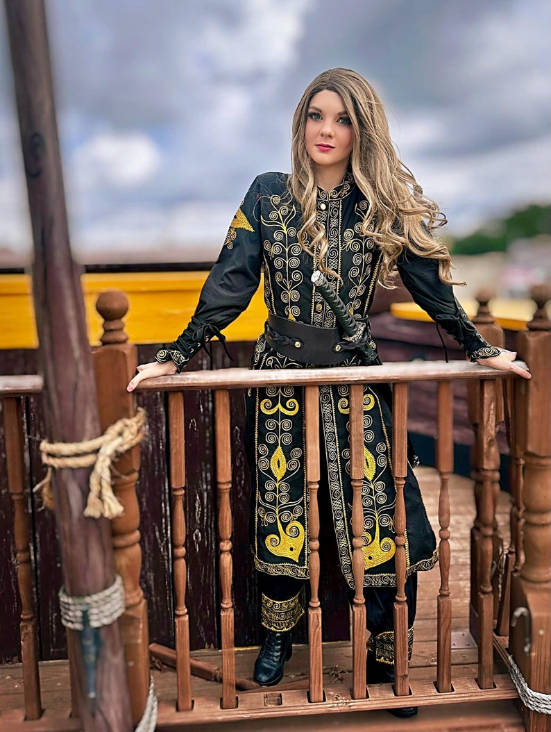 A woman in a black and gold embroidered coat stands on a wooden deck. She is holding a sword in her right hand and looking off to the side. The coat has a high collar and long sleeves. The embroidery is intricate and detailed. The woman's hair is long and blonde. She is wearing black pants and black boots. Elizabeth Swann costume from pirates of the Caribbean
