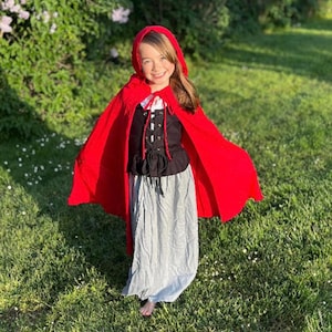 May include: A child's costume featuring a bright red hooded cape, a black bodice with a laced front, and a long, light gray skirt. The costume is designed for dress-up and imaginative play, perfect for Halloween or themed events.