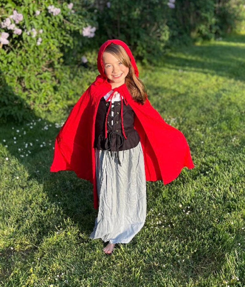 May include: A child's costume featuring a bright red hooded cape, a black bodice with a laced front, and a long, light gray skirt. The costume is designed for dress-up and imaginative play, perfect for Halloween or themed events.