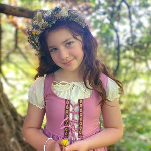 A young girl wearing a pink dress with a floral crown and holding a yellow flower.