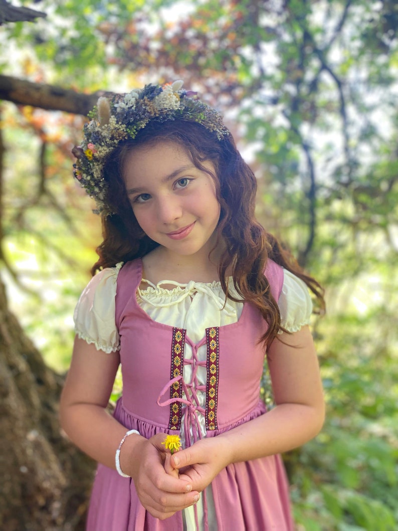 A young girl wearing a pink dress with a floral crown and holding a yellow flower.