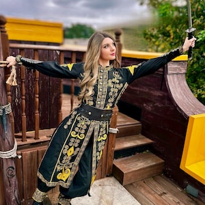A woman in a black and gold embroidered costume from pirates of the Caribbean holds a sword in her right hand. She is standing on a wooden platform with a yellow railing.