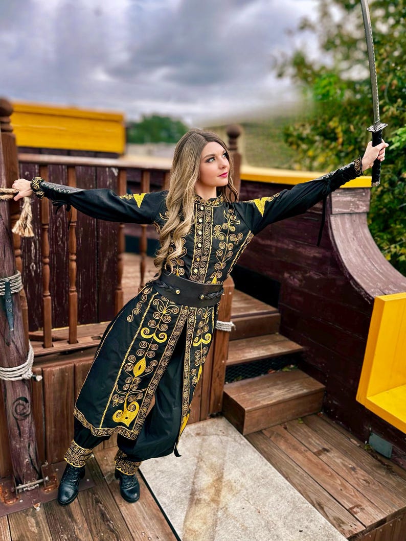 A woman in a black and gold embroidered costume from pirates of the Caribbean holds a sword in her right hand. She is standing on a wooden platform with a yellow railing.