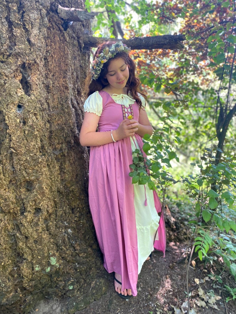 A young girl wearing a pink dress and white under dress with a floral crown stands in a forest setting. She is holding a small flower in her hand.