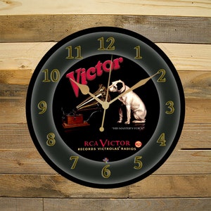 May include: A black clock with gold numbers and hands. The clock face features a vintage RCA Victor logo with a dog listening to a phonograph. The text "His Master's Voice" and "RCA Victor Records Victrolas Radios" is printed on the clock face.