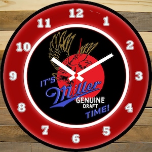 May include: A round wall clock with a red outer ring and a black center. The clock face features a red circle with a gold eagle graphic and the words "IT'S Miller GENUINE DRAFT TIME!" in white and blue. White hands indicate the time.