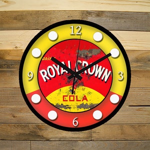 May include: A round clock with a black frame and white numbers. The clock face features a vintage Royal Crown Cola logo with red, yellow, and white colors. The logo reads "Drink Royal Crown Cola".