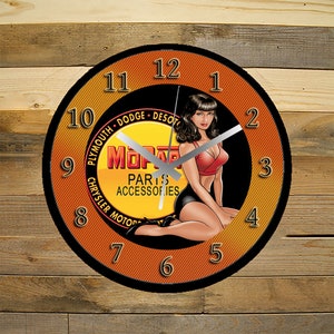 May include: A round orange and black clock with a pin-up girl illustration. The clock face features the text "Mopar Parts Accessories" and the names of car brands: "Plymouth, Dodge, DeSoto, Chrysler Motors".
