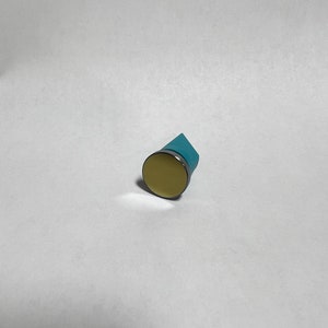 May include: A small, round, light yellow object with a silver rim and a blue backing. The object is set against a white background. The item appears to be a small magnet.