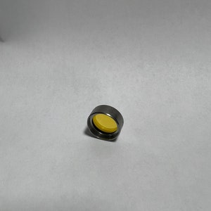 Pode incluir: Um pequeno objeto cilíndrico metálico com uma lente amarela e circular. O objeto é prateado e parece ser feito de metal. A lente amarela é visível dentro do cilindro.