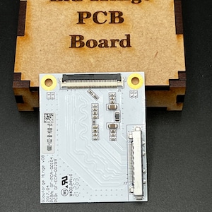Puede incluir: Una placa de circuito impreso blanca con un conector negro y el texto "Glowforge Hinge v08" impreso en ella. La placa tiene un conector negro en la parte superior y un conector blanco en la parte inferior. La placa está etiquetada con el texto "PCBA: GF-PCA-00104" y "PCB: GF-ECA-00299".