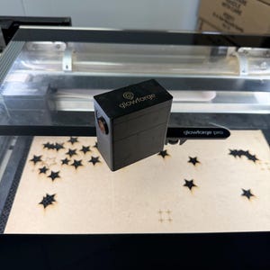 May include: A black Glowforge Pro laser cutter with the brand name on top. The machine is cutting star shapes from a light brown material. The machine has a clear lid and a light inside.