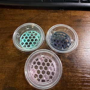 May include: Three circular clear plastic containers, each featuring a removable lid with a built-in hexagonal grid insert. The inserts are colored light mint green, dark navy blue, and translucent light pink, respectively, for organizing small items.