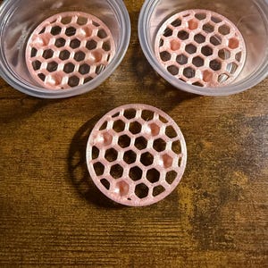 May include: Two clear plastic circular containers, each containing a light pink, circular, 3D-printed insert with a hexagonal honeycomb pattern. A third identical light pink honeycomb insert is positioned separately below the two containers.
