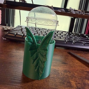 May include: A clear plastic cup with a domed lid inserted into a cylindrical green holder. The holder features a prominent, textured, three-dimensional green leaf design attached to its exterior, extending vertically along the side of the cup.