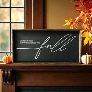 May include: A rectangular wooden sign with a black background and a dark wood frame. The sign features white text that reads, "and all at once summer collapsed into fall," with the word "fall" written in a large, flowing, cursive script.