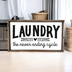 K&ouml;nnte beinhalten: Ein Holzschild mit dem Text "Laundry the never ending cycle" in schwarzen Buchstaben. Das Schild hat einen rustikalen braunen Rahmen und einen wei&szlig;en Hintergrund.
