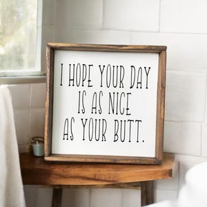 May include: A framed wooden sign with the text "I HOPE YOUR DAY IS AS NICE AS YOUR BUTT" in black lettering. The sign is displayed on a wooden table in a bathroom setting.