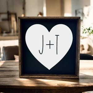 May include: A wooden framed sign with a black background and a white heart shape. The letters "J + T" are written in black inside the heart. The sign is displayed on a wooden table.