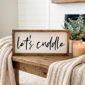 May include: A rectangular wooden sign with the words "let's cuddle" in black script. The sign is set on a wooden table with a cream-colored blanket draped over it. A lit candle and a potted plant are also on the table.