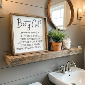 May include: A wooden framed sign with the words "Booty Call" in black script. The sign includes a definition and is displayed on a rustic wooden shelf with a small potted plant, a glass jar, and a round mirror.