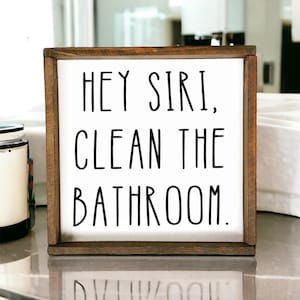 May include: A framed wooden sign with the text "HEY SIRI, CLEAN THE BATHROOM." The sign has a white background and black lettering. The frame is a dark brown color. The sign is a decorative item for a bathroom.