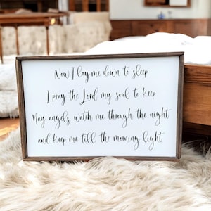 May include: A white wooden sign with black text that reads "Now I lay me down to sleep I pray the Lord my soul to keep May angels watch me through the night and keep me till the morning light".
