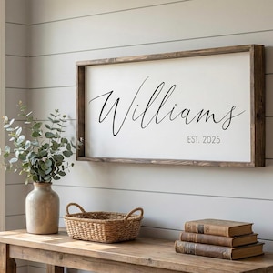 May include: A framed wooden sign with the name "Williams" in elegant script and the date "EST. 2025". The sign is displayed above a wooden table with a vase, basket, and books. The wall is painted light gray.