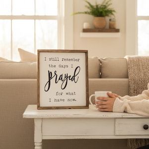 May include: A framed wooden sign with the text "I still remember the days I prayed for what I have now." The sign is on a white table, and a person is holding a white mug. The background includes a couch and a window.