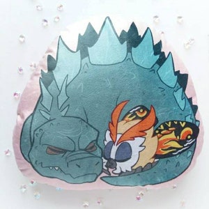 May include: A round, plush pillow featuring a cartoon illustration of Godzilla and Mothra. Godzilla is a teal-green color with spiky dorsal fins, and Mothra is depicted with orange, white, and black wings. The pillow has a pink border.