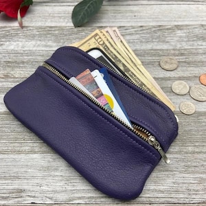 May include: A dark purple leather coin purse with a silver zipper, open to reveal cash, credit cards, and a smartphone. The purse is on a wooden surface, with loose change scattered nearby.