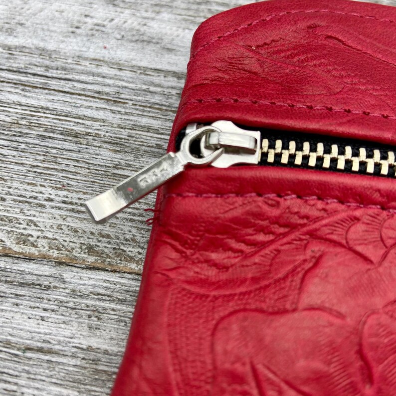 May include: Close-up of a red leather wallet with a silver zipper. The leather has a floral embossed design. The zipper is open, revealing a black interior.