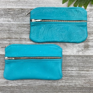 May include: Two turquoise leather pouches with silver zippers. One pouch features an embossed floral design, while the other is smooth. Both have a rounded rectangular shape, perfect for carrying small items.