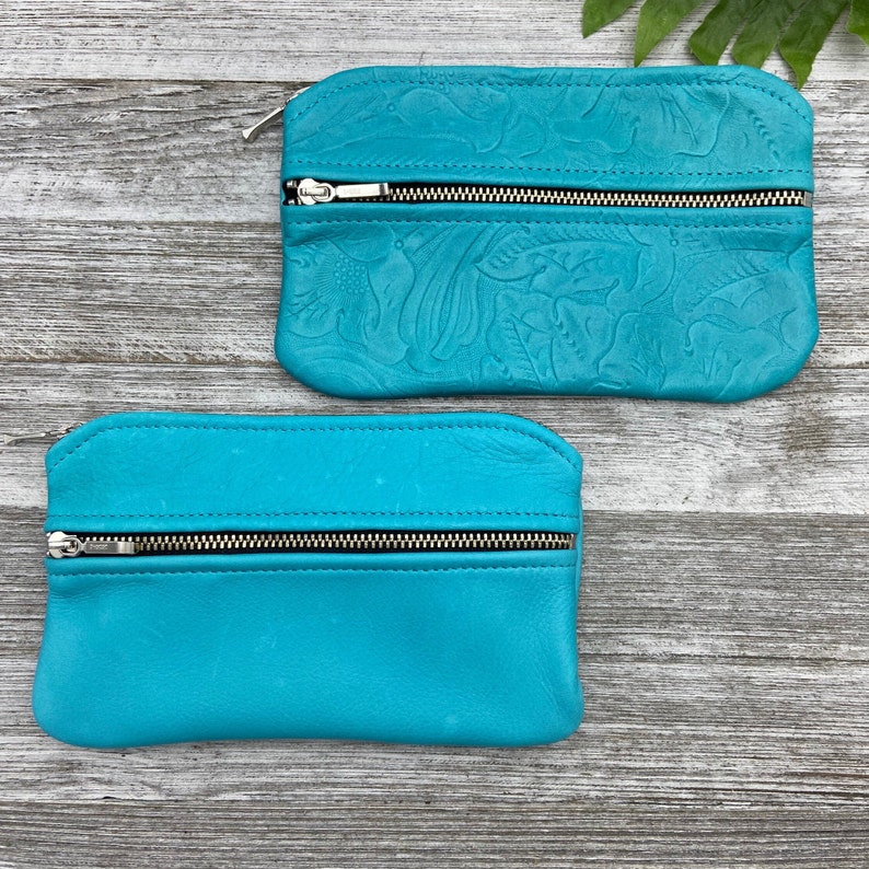 May include: Two turquoise leather pouches with silver zippers. One pouch features an embossed floral design, while the other is smooth. Both have a rounded rectangular shape, perfect for carrying small items.