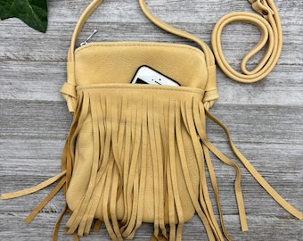 May include: A tan leather crossbody bag with a long strap and fringe detail. A smartphone is visible in the front pocket. The bag is made of soft leather and has a zipper closure. The bag is on a wooden surface.