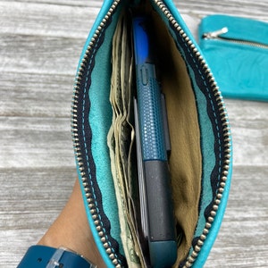 May include: A turquoise leather zippered pouch, open to reveal cash and a blue phone. The interior is tan, with black stitching and a silver zipper. A matching pouch is visible in the background.