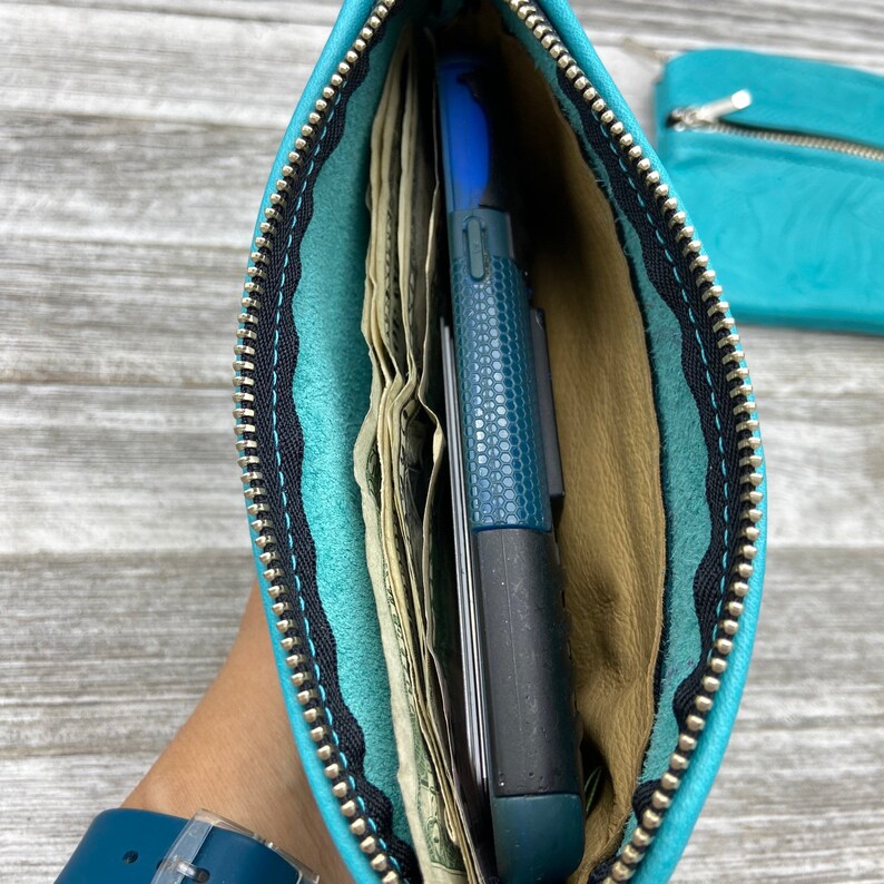 May include: A turquoise leather zippered pouch, open to reveal cash and a blue phone. The interior is tan, with black stitching and a silver zipper. A matching pouch is visible in the background.
